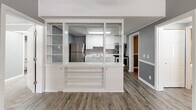 Located at 1300 Nashville Pike in Gallatin, Tennessee, this two-bedroom, two-bathroom unit offers a practical and well-organized single-floor layout. The kitchen is a standout feature, defined by white cabinetry, white countertops, stainless steel appliances including a refrigerator, range, and microwave, and a stainless steel sink. A distinctive built-in shelving unit with open shelves and glass panels separates the kitchen from the living area, adding architectural character while maintaining an open feel. Hardwood-style flooring runs through the kitchen, hallway, and laundry areas, lending a cohesive look throughout the common spaces. Both bedrooms are finished with plush carpeting and feature ceiling fans, natural light from windows with blinds, and a two-tone gray wall treatment with white chair rail molding. The spacious primary bedroom includes its own en-suite bathroom, which offers a white vanity with ample cabinet storage and a separate toilet area. The second bathroom features a full tub-shower combo and matching white vanity. A dedicated laundry room accommodates a full-size washer and dryer, adding everyday convenience. The textured ceilings and consistent gray-toned palette throughout give the unit a clean, uniform aesthetic.