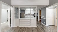 Located at 1300 Nashville Pike in Gallatin, Tennessee, this two-bedroom, two-bathroom unit offers a practical and well-organized single-floor layout. The kitchen is a standout feature, defined by white cabinetry, white countertops, stainless steel appliances including a refrigerator, range, and microwave, and a stainless steel sink. A distinctive built-in shelving unit with open shelves and glass panels separates the kitchen from the living area, adding architectural character while maintaining an open feel. Hardwood-style flooring runs through the kitchen, hallway, and laundry areas, lending a cohesive look throughout the common spaces. Both bedrooms are finished with plush carpeting and feature ceiling fans, natural light from windows with blinds, and a two-tone gray wall treatment with white chair rail molding. The spacious primary bedroom includes its own en-suite bathroom, which offers a white vanity with ample cabinet storage and a separate toilet area. The second bathroom features a full tub-shower combo and matching white vanity. A dedicated laundry room accommodates a full-size washer and dryer, adding everyday convenience. The textured ceilings and consistent gray-toned palette throughout give the unit a clean, uniform aesthetic.