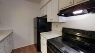 This single-floor residence offers a practical and well-organized living space. The kitchen is a standout feature, designed for efficient use with a clean, classic aesthetic. White upper and lower cabinetry provides ample storage, complemented by a crisp white countertop that offers generous prep space. Black appliances, including a refrigerator, electric range with a smooth cooktop, and a range hood above, create a bold contrast against the light cabinetry and walls. The range hood is neatly integrated beneath the upper cabinets, contributing to a streamlined look. The kitchen layout makes efficient use of the available space, positioning the refrigerator and range in close proximity for a practical workflow. Warm hardwood flooring runs through the space, adding a touch of natural texture and warmth to the interior. The walls are finished in a neutral, light tone that keeps the space feeling bright and open. An electrical panel is conveniently located within the kitchen area, and standard electrical outlets are accessible along the walls. Overall, the space presents a straightforward, functional design with durable finishes suited for everyday living.