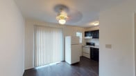 This multi-level home spans three floors, offering a well-organized layout with distinct living zones across each level. The main floor features dark hardwood-style flooring that flows through the open living area and into the kitchen. The kitchen is appointed with dark cabinetry, white tile backsplash, a full suite of appliances including a refrigerator, electric range, and dishwasher, along with a sink set into light countertops. A ceiling fan and large sliding glass door with vertical blinds fill the adjacent living space with natural light. A carpeted staircase with a sleek dark metal railing connects the floors. The upper level hosts carpeted bedrooms, one of which features a large window with outdoor views and generous closet space with dark wood sliding doors. The bathroom includes a dark vanity with a white countertop, tile-surround soaking tub with shower, and hard surface flooring. The lower level provides an expansive unfinished basement with a painted concrete floor, sump pump, electrical panel, and a small egress window — offering significant potential for additional storage or future finishing. Throughout the home, neutral white walls and consistent finishes create a clean, cohesive atmosphere.