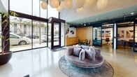Located at 3800 Mentone Ave in Culver City, California, this single-floor lobby space combines industrial and contemporary design elements to create a welcoming, functional environment. The interior is defined by soaring ceilings with exposed concrete columns and visible ductwork, lending an open, loft-like character throughout.

The entry area features floor-to-ceiling glass walls and a wide glass door that flood the space with abundant natural light and provide clear views to the surrounding streetscape. Polished concrete flooring extends across the entire footprint, reinforcing the modern industrial aesthetic.

At the heart of the lobby sits a circular, velvet sofa in a soft lavender hue, centered on a round area rug—serving as a comfortable, visually distinctive lounge focal point. Overhead, a dramatic cluster of oversized white globe pendant lights adds warmth and a sculptural quality to the high ceiling. Decorative wall-mounted medallions provide additional artistic interest.

A casual dining area with a round wood table and black spindle chairs is positioned near the glazed perimeter, accompanied by a built-in wood credenza. A bookshelf-lined reading nook offers a quieter retreat. Two stainless steel elevators are accessible from the lobby, with exposed concrete walls nearby reinforcing the building's architectural identity.