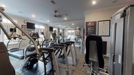 Located at 5200 Greystone Summit Dr in Columbus, Georgia, this fitness center combines functional design with refined architectural details to create a welcoming and well-appointed workout environment. The single-floor space is finished with crown molding, recessed lighting, and neutral warm-toned walls that lend a polished, upscale feel throughout.

Natural light pours in through multiple multi-pane windows positioned along several walls, keeping the space bright and open. A ceiling fan provides additional climate comfort, while large wall-mounted mirrors visually expand the room and allow users to monitor their form during exercise.

The cardio section is comprehensively outfitted with treadmills — including Star Trac and Cybex models — elliptical trainers, stationary bikes, and stair-climbing machines, several of which feature personal viewing screens. The strength training area includes a cable and pulley multi-station machine, a Cybex weight machine, and a full dumbbell rack. Stability and exercise balls are neatly organized on a dedicated rack, rounding out the functional fitness options.

Patterned carpet tiles cover much of the floor, complemented by rubber mat sections beneath the equipment. Framed informational fitness posters and posted fitness center rules add a practical, community-oriented touch to the space, making it a thorough and thoughtfully designed amenity.