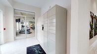 This single-floor amenity space is designed with a clean, modern aesthetic that prioritizes both function and visual appeal. The corridor serves as the central hub connecting key community amenities, finished throughout with light-toned large-format tile flooring that enhances the bright, airy atmosphere.

Prominently lining one wall is a floor-to-ceiling bank of package lockers, offering a secure and convenient parcel retrieval system. Adjacent to the lockers, a built-in water fountain provides a practical touch for residents on the go.

Glass-paneled double doors open into a well-appointed fitness center, visible from the corridor and flooded with natural light. The gym is equipped with treadmills, a ceiling fan, wall-mounted television, resistance training equipment, and exercise balls, making it a comprehensive space for a variety of workouts.

Further along the corridor, a second set of glass double doors leads to what appears to be a conference or meeting room, featuring a large table, task chairs, and decorative greenery. Built-in wall shelving with decorative wooden accents adds warmth and character to the connecting hallway.

Throughout, the white walls, recessed lighting, and consistent flooring create a cohesive, polished environment that reflects a high standard of community amenity design.