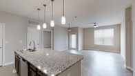 This contemporary single-floor residence showcases a thoughtfully designed open-concept layout that seamlessly connects the kitchen, dining, and living areas. The striking herringbone-patterned luxury vinyl flooring flows consistently throughout, lending a cohesive and sophisticated aesthetic to the entire space. Neutral warm-gray walls complement the clean white trim and doors found in every room.

The kitchen serves as the centerpiece, featuring rich dark-espresso cabinetry, granite countertops with subtle veining, a white subway tile backsplash, and a full suite of stainless steel appliances including a refrigerator, range, and built-in microwave. A generously sized kitchen island with an undermount sink provides additional prep space and seating potential, illuminated by a series of modern pendant lights and supplemented by track lighting overhead.

The living and dining areas benefit from multiple windows with horizontal blinds, allowing ample natural light. A ceiling fan adds comfort to the main living space, while a pendant light fixture anchors the dining area near the entry door.

The primary bathroom offers a long, extended vanity with dark cabinetry, a backlit mirror, and ample storage. A full tub-shower combination with white tile surround is found in the secondary bathroom alongside matching dark cabinetry above. The bedrooms are bright, benefiting from large windows that invite natural light.