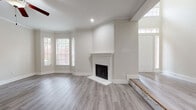 This two-story home showcases a clean, modern aesthetic with neutral tones and thoughtful finishes throughout. The grand foyer greets guests with a soaring transom-lit entry door and elegant marble-accented steps that lead down into the expansive main living area. The living room is anchored by a classic white-mantled fireplace and features light gray hardwood-style flooring, crown molding, and a bay window that draws in generous natural light. The kitchen is outfitted with crisp white cabinetry, stainless steel appliances including a refrigerator, range, and over-the-range microwave, all set atop matching light wood-style flooring. A separate dining or flex room with crown molding and a ceiling fan offers additional versatile living space. Upstairs, an open landing with a half-wall overlook connects the upper-level rooms. The spacious primary bedroom features plush carpet, a bay window, vaulted ceiling, and a ceiling fan. The luxurious en-suite bathroom includes a large walk-in glass shower with a rainfall showerhead and mosaic tile niche, a dual-sink vanity with dark granite countertops, and an expansive walk-in closet. A secondary bathroom features a soaking tub with tiled surround, a mosaic niche, and a dark granite vanity.