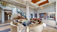 Located at 4015 Aspen Grove Dr in Franklin, TN, this impressive community clubhouse delivers a sophisticated, hotel-inspired atmosphere across a single, expansive floor. The space is immediately striking upon entry, where double-height ceilings and floor-to-ceiling windows flood the interior with natural light and offer sweeping views of the surrounding landscape. The entry vestibule features a refined mix of marble-look tile and herringbone-patterned flooring, with a decorative chevron-tiled accent wall adding visual interest near the double-door entrance.

The main gathering area is anchored by a dramatic open-plan layout, with distinct seating and dining zones furnished with marble-topped round tables and mid-century modern wooden chairs. A central bar and kitchen station—clad in mosaic tile and outfitted with pendant lighting—serves as a social hub, complemented by bar-height seating along its perimeter. A grand staircase with glass railings ascends to a mezzanine level featuring ornamental screen panels, adding architectural depth to the space.

Warm wood ceiling panels with statement chandeliers contrast beautifully with the cool-toned tile floors throughout. Lush tropical plants are strategically positioned to bring life and greenery indoors. The overall ambiance is contemporary and upscale, blending open communal spaces with intimate seating nooks, all bathed in abundant natural light.