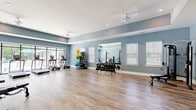 Located at 4605 Sidbury Crossing Dr in Castle Hayne, NC, this fitness center offers a thoughtfully designed exercise space across a single, open floor. The room is finished with durable wood-look flooring and painted in a calming steel-blue palette, complemented by crisp white trim and a tray ceiling detail that adds architectural dimension. Ceiling fans provide consistent air circulation throughout the space.

Natural light is a defining feature, with multiple windows fitted with white blinds lining the walls, and a dramatic floor-to-ceiling glass window wall that looks directly out onto a resort-style outdoor pool and lush tree line — creating an inviting, energizing atmosphere during workouts.

The fitness center is comprehensively equipped for a full range of training modalities. Cardio equipment includes treadmills, an elliptical, and a stationary bike, positioned to take advantage of the pool views. The free weight section features a full dumbbell rack and stability balls on a dedicated storage stand. Strength training is supported by a full power rack with a bench press setup and a cable machine. A wall-mounted television and a pull-up/dip tower round out the amenities. A large mirror on one wall enhances the sense of space. French doors connect the fitness room to an adjacent common area.