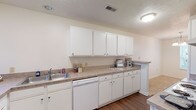 This single-story unit at The Brickyard offers a thoughtfully arranged layout with a clean, neutral aesthetic throughout. The kitchen serves as a functional centerpiece, featuring an expansive run of white cabinetry — both upper and lower — paired with laminate countertops that provide ample workspace. White appliances, including a full-size refrigerator, dishwasher, and range, are seamlessly integrated into the design. Dark hardwood-style flooring runs through the kitchen, adding warmth and contrast to the bright cabinetry. A window above the sink area allows natural light to filter in.

The living area transitions to plush carpeting and is highlighted by a ceiling fan and a large sliding glass door fitted with vertical blinds, offering views of lush outdoor greenery and providing access to the exterior. An adjacent window further enhances natural light in the space.

The bedrooms are generously sized, each finished with soft carpeting, ceiling fans with light fixtures, and windows that bring in natural daylight. Closet storage is included in the bedroom areas.

The bathroom features a white vanity with a large mirror, well-lit by a Hollywood-style light bar above. Dark hardwood-style flooring continues here, complementing the clean white fixtures and a shower-tub combination.