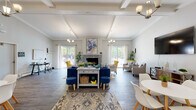 This thoughtfully designed single-floor space makes a strong first impression, accessible through elegant French doors that open into a expansive great room. The vaulted, beam-accented ceiling soars overhead, lending the space an airy, open quality filled with natural light streaming in from multiple sliding glass doors on opposing walls — offering views of the surrounding outdoor areas.

At the heart of the great room stands a stacked stone fireplace topped with an eye-catching abstract art piece, flanked by vibrant yellow accent chairs and a navy blue sofa that add bold pops of color against the neutral gray walls and dark hardwood-style flooring. A central console table anchors the living area, while a dedicated seating vignette with taupe armchairs and a mounted flat-screen television creates a comfortable media zone along one wall.

Two elegant chandeliers provide warm, ambient lighting throughout the main living space, complementing the natural light throughout the day. A dining area with a round white table is positioned near the entrance, adding functional versatility to the open floor plan.

The kitchen features crisp white shaker cabinetry, dark granite countertops, and stainless steel appliances, delivering a clean and classic aesthetic. Throughout, the space balances modern comfort with warm, inviting design details, creating a welcoming and highly functional environment.