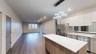 This well-appointed 3-bedroom, 2-bathroom residence is thoughtfully designed on a single floor, offering a seamless and functional layout. The open-concept living and kitchen area forms the heart of the home, anchored by a striking kitchen that features a large center island with a white quartz countertop, dual-tone cabinetry with warm wood lower cabinets and crisp white upper cabinets, a mirrored backsplash, stainless steel appliances including a French door refrigerator and range, a wall-mounted range hood, and a modern cluster pendant light fixture overhead. The expansive living area flows naturally from the kitchen, bathed in natural light from large windows and sliding glass doors that offer views of the surrounding neighborhood. Hardwood-style flooring runs consistently throughout, adding warmth and cohesion to the space. The generous primary bedroom provides a comfortable retreat, while the two additional bedrooms offer flexibility for guests or a home office, each with hardwood-style floors and good natural light. Both bathrooms are finished with wood-toned vanities, white countertops, and clean, neutral tile work. The home is completed by a utility room, a closet for additional storage, and recessed lighting throughout, resulting in a bright, contemporary living environment.
