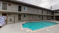 This two-story residential complex is centered around a generously sized outdoor courtyard that serves as the social hub of the property. The courtyard features a large, vivid turquoise swimming pool with a smooth concrete deck providing ample space for lounging and recreation. The pool area is enclosed by wrought iron fencing, ensuring both safety and an open, airy feel. Overhead, decorative string lights are strung across the courtyard, adding a warm ambiance during evening hours.

The building's exterior is finished in a textured stucco facade, with units on both the ground and upper levels opening onto private balconies or patios equipped with iron railings. Large windows throughout allow natural light to fill interior spaces and offer views of the pool and courtyard below. A bold red accent wall on one wing of the building adds a striking visual contrast to the neutral tones of the overall structure.

A tall palm tree rises from the central courtyard landscaping, contributing to the property's warm, Southwestern character. An on-site fitness center, open daily from 8 AM to 8 PM, provides residents with a convenient exercise facility. The combination of resort-style outdoor amenities, thoughtful architectural details, and communal spaces makes this property a comfortable and inviting place to call home.