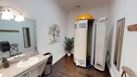 This tanning room is a well-appointed, single-level space designed to provide a comfortable and functional tanning experience. The centerpiece of the room is a stand-up tanning unit with a distinctive yellow dome top and a navy blue base, complemented by an open panel unit positioned nearby, offering users multiple tanning options.

The room features rich, dark hardwood-style flooring that contrasts cleanly with the bright white walls accented by classic crown molding. A granite-topped vanity with a single basin sink and a wall-mounted light fixture provides a practical grooming station, accompanied by a large mirror. A full-length mirror with a decorative silver frame offers an additional spot to check results post-session.

Thoughtful touches include a rustic wooden wall-mounted coat hook rack and a folding stool, providing convenient space for personal items. A potted palm plant adds a natural, welcoming accent, while a colorful watercolor tree painting contributes a polished, salon-like aesthetic. A six-panel door with brushed nickel hardware provides private entry, completing this clean and practical tanning environment.