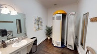 This tanning room is a well-appointed, single-level space designed to provide a comfortable and functional tanning experience. The centerpiece of the room is a stand-up tanning unit with a distinctive yellow dome top and a navy blue base, complemented by an open panel unit positioned nearby, offering users multiple tanning options.

The room features rich, dark hardwood-style flooring that contrasts cleanly with the bright white walls accented by classic crown molding. A granite-topped vanity with a single basin sink and a wall-mounted light fixture provides a practical grooming station, accompanied by a large mirror. A full-length mirror with a decorative silver frame offers an additional spot to check results post-session.

Thoughtful touches include a rustic wooden wall-mounted coat hook rack and a folding stool, providing convenient space for personal items. A potted palm plant adds a natural, welcoming accent, while a colorful watercolor tree painting contributes a polished, salon-like aesthetic. A six-panel door with brushed nickel hardware provides private entry, completing this clean and practical tanning environment.