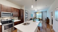 This well-appointed one-bedroom, one-bathroom residence at 1201 Ashford Cir in Fredericksburg, Virginia offers a thoughtfully designed open-concept floor plan across a single level. The kitchen is a standout feature, showcasing rich dark-wood cabinetry, a mosaic tile backsplash, stainless steel appliances, and a generous white quartz island with a built-in sink and pendant lighting — flowing seamlessly into the dining area and living room. The dining space accommodates a full table beneath a stylish drum pendant light, while warm hardwood-style flooring runs throughout the main living areas. The living room is bright and welcoming, with large windows dressed in patterned drapes and blinds that invite natural light, complemented by a ceiling fan overhead. The spacious primary bedroom features plush carpeting, large windows with ample natural light, a ceiling fan, and direct access to both a walk-in closet and the full bathroom. The bathroom impresses with a dual-sink vanity topped in white quartz, dark wood cabinetry, and well-lit mirrors. A dedicated closet provides additional storage. The overall ambiance is modern yet comfortable, with a cohesive neutral palette and upscale finishes throughout.