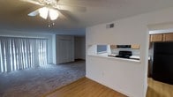 This single-floor residence offers a practical and open layout that seamlessly connects the main living areas. The living room is finished with plush wall-to-wall carpeting and features a ceiling fan with a light fixture overhead, providing both comfort and air circulation. A textured ceiling adds a classic touch throughout the space. Natural light filters in through vertical blinds framing a sliding glass door, which leads to an outdoor area and brightens the interior. A pass-through counter connects the living area to the kitchen, creating an open and communicative flow between the two spaces. The kitchen is equipped with a black electric range, a built-in microwave, and light wood cabinetry, offering functional storage and prep space. The transition between the kitchen and living areas is marked by a shift from hardwood-style flooring to carpet, clearly delineating each zone. A closet with bi-fold doors is visible from the main living area, providing convenient storage. The walls are painted in a clean, neutral white, contributing to a bright and airy atmosphere. The overall layout is efficient and well-organized, making excellent use of the available floor plan on a single level.