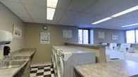 This shared laundry facility is designed with functionality and practicality in mind. The room is equipped with a row of front-load washing machines, positioned along a central island topped with granite-style countertops that provide ample space for folding and sorting laundry. Additional countertop space lines the walls, further enhancing usability for multiple users at once.

The flooring features a classic black-and-white checkered tile pattern, adding a timeless aesthetic contrast to the otherwise neutral, utilitarian design. The walls are painted in a warm beige tone, complemented by a suspended ceiling fitted with bright fluorescent panel lighting that ensures the space is well-lit throughout.

A window along the back wall allows natural light to enter and offers a view of the surrounding urban environment. Informational notices and usage instructions are posted on the walls, providing clear guidance for users. A wall clock keeps residents on schedule during their laundry sessions. The space is organized, clean, and efficiently laid out to accommodate the needs of multiple residents in a shared building setting.