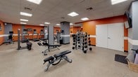 This fitness center is a comprehensively outfitted single-floor facility designed to support a full range of workout routines. The space is defined by its bold color scheme, featuring vibrant orange accent walls contrasted with gray columns and a crisp white horizontal stripe that runs throughout, creating an energetic and motivating atmosphere. A drop ceiling with recessed panel lighting provides consistent, even illumination across the entire floor area.

The cardio zone lines one wall and houses multiple treadmills and elliptical machines, each equipped with individual monitors for entertainment during workouts. Adjacent to this area, the free weights section offers a well-stocked dumbbell rack with a full range of weights, kettlebells, and stability balls, accompanied by adjustable benches for a variety of strength exercises.

Cable and resistance machines are strategically positioned throughout the space, offering versatile options for both upper and lower body training. A dedicated stretching and flexibility area features foam mats stored in a wicker basket, interlocking floor padding, and a wall-mounted television for guided sessions.

Full-length mirrors line multiple walls, amplifying the sense of space and allowing users to monitor their form. The durable speckled flooring is both practical and easy to maintain. The open layout of the facility ensures ample room to move freely between equipment zones.