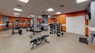 This fitness center is a comprehensively outfitted single-floor facility designed to support a full range of workout routines. The space is defined by its bold color scheme, featuring vibrant orange accent walls contrasted with gray columns and a crisp white horizontal stripe that runs throughout, creating an energetic and motivating atmosphere. A drop ceiling with recessed panel lighting provides consistent, even illumination across the entire floor area.

The cardio zone lines one wall and houses multiple treadmills and elliptical machines, each equipped with individual monitors for entertainment during workouts. Adjacent to this area, the free weights section offers a well-stocked dumbbell rack with a full range of weights, kettlebells, and stability balls, accompanied by adjustable benches for a variety of strength exercises.

Cable and resistance machines are strategically positioned throughout the space, offering versatile options for both upper and lower body training. A dedicated stretching and flexibility area features foam mats stored in a wicker basket, interlocking floor padding, and a wall-mounted television for guided sessions.

Full-length mirrors line multiple walls, amplifying the sense of space and allowing users to monitor their form. The durable speckled flooring is both practical and easy to maintain. The open layout of the facility ensures ample room to move freely between equipment zones.
