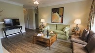 This single-floor residence offers a welcoming and thoughtfully designed interior highlighted by rich, dark hardwood-style flooring that runs throughout the main living area, adding warmth and visual continuity to the space. The living room serves as the central gathering area, featuring crown molding along the ceiling and a ceiling fan with integrated lighting that provides both comfort and illumination. A glass-topped wooden coffee table anchors the seating arrangement, complemented by a sage green sofa dressed with accent pillows and flanked by side tables with warm-toned lamps, creating an inviting and well-balanced ambiance. A framed abstract artwork in earth tones adds a decorative focal point above the sofa, enhancing the room's character. The open layout allows sightlines into the adjoining hallway, which leads to additional rooms beyond, suggesting a functional and connected floor plan. White panel doors and neutral-toned walls provide a clean backdrop throughout, while the overall design balances classic furnishings with practical living elements, making the space feel both comfortable and tastefully appointed.