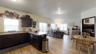 This single-floor home is designed around an open-concept layout that effortlessly connects the kitchen, dining, and living areas into one cohesive, flowing space. The kitchen features dark cabinetry that provides a striking contrast against the light-toned hardwood flooring found throughout the home. A peninsula with counter seating serves as both a functional workspace and a natural dividing point between the kitchen and the rest of the living space, offering convenient casual dining. The dining area accommodates a full table and chairs, positioned to take advantage of the natural light streaming in through multiple large windows. The living room is generously sized, offering ample room for comfortable seating arrangements, and benefits from the same bright, airy atmosphere created by the large windows along the exterior walls. The continuity of the light hardwood flooring across all areas enhances the sense of openness and flow throughout the home. Neutral wall tones complement the overall design, creating a warm and welcoming ambiance. The single-floor layout ensures easy accessibility and a practical, connected flow between all living spaces, making it a functional and comfortable home.