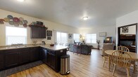 This single-floor home is designed around an open-concept layout that effortlessly connects the kitchen, dining, and living areas into one cohesive, flowing space. The kitchen features dark cabinetry that provides a striking contrast against the light-toned hardwood flooring found throughout the home. A peninsula with counter seating serves as both a functional workspace and a natural dividing point between the kitchen and the rest of the living space, offering convenient casual dining. The dining area accommodates a full table and chairs, positioned to take advantage of the natural light streaming in through multiple large windows. The living room is generously sized, offering ample room for comfortable seating arrangements, and benefits from the same bright, airy atmosphere created by the large windows along the exterior walls. The continuity of the light hardwood flooring across all areas enhances the sense of openness and flow throughout the home. Neutral wall tones complement the overall design, creating a warm and welcoming ambiance. The single-floor layout ensures easy accessibility and a practical, connected flow between all living spaces, making it a functional and comfortable home.
