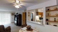 This single-floor residence features an open, functional layout that promotes a sense of connected living. The dining area serves as a central hub, furnished with a round table beneath a classic ceiling fan with integrated lighting, which provides both airflow and ambient illumination. Soft carpeting underfoot adds warmth to the space.

A defining architectural feature is the wide pass-through window cut into the wall between the dining area and kitchen, allowing natural sightlines and easy communication between the two spaces. The kitchen is well-appointed with warm wood-toned upper cabinetry, a white electric range, and an over-the-range microwave. A built-in range hood and dark microwave/oven unit contribute to a practical cooking environment.

Adjacent to the dining space, a dark wood entertainment unit provides media storage, while a built-in shelving unit and interior door hint at additional storage or closet space nearby. Framed artwork on the walls adds a personal touch to the neutral color palette. The overall ambiance is cozy and practical, with a straightforward layout designed for comfortable everyday living.
