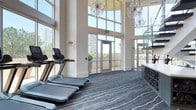 This upscale two-story fitness center seamlessly blends elegant design with high-performance functionality. The main floor is anchored by dramatic double-height ceilings wrapped in expansive floor-to-ceiling glass curtain walls, flooding the space with natural light and offering sweeping views of the surrounding cityscape and greenery. Ornate crystal chandeliers suspended from above lend an unexpected touch of grandeur, creating a striking contrast against the sleek, modern architecture. A glass-railed staircase connects the two levels with visual openness throughout.

The lower level houses a well-equipped strength and cardio zone, featuring treadmills, stationary bikes, a cable machine, dumbbell racks with adjustable benches, and a dedicated functional training station complete with a punching bag, TRX suspension system, and plyosoft boxes. Large wall-mounted mirrors expand the sense of space across the workout areas.

The upper level offers a dedicated studio space outfitted with wood-tone built-in shelving stocked with fitness accessories, yoga mats, and step platforms — ideal for group classes or individual training sessions. Glass-paneled sliding doors provide flexible room division. Throughout the facility, hardwood-style and patterned carpet flooring define distinct zones, while recessed lighting and clean white walls maintain a bright, polished atmosphere.