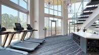 This upscale two-story fitness center seamlessly blends elegant design with high-performance functionality. The main floor is anchored by dramatic double-height ceilings wrapped in expansive floor-to-ceiling glass curtain walls, flooding the space with natural light and offering sweeping views of the surrounding cityscape and greenery. Ornate crystal chandeliers suspended from above lend an unexpected touch of grandeur, creating a striking contrast against the sleek, modern architecture. A glass-railed staircase connects the two levels with visual openness throughout.

The lower level houses a well-equipped strength and cardio zone, featuring treadmills, stationary bikes, a cable machine, dumbbell racks with adjustable benches, and a dedicated functional training station complete with a punching bag, TRX suspension system, and plyosoft boxes. Large wall-mounted mirrors expand the sense of space across the workout areas.

The upper level offers a dedicated studio space outfitted with wood-tone built-in shelving stocked with fitness accessories, yoga mats, and step platforms — ideal for group classes or individual training sessions. Glass-paneled sliding doors provide flexible room division. Throughout the facility, hardwood-style and patterned carpet flooring define distinct zones, while recessed lighting and clean white walls maintain a bright, polished atmosphere.
