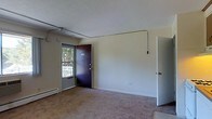 This single-floor space presents a straightforward and functional layout centered around a carpeted main living area. The room is finished with neutral-toned walls and a textured ceiling, contributing to a clean, understated aesthetic. An exposed metal conduit runs along the ceiling and upper wall, adding an utilitarian architectural detail to the space.

The entry area features a bold-colored front door with dual locks, accompanied by a side window that allows natural light to filter into the room while offering a view of an outdoor railing and greenery beyond. A baseboard heater sits beneath the window, providing a practical heating solution along the exterior wall.

The carpeting throughout the main area is a muted, neutral tone, lending a soft underfoot feel to the living space. An additional interior door is visible along the adjacent wall, suggesting access to further rooms or storage within the unit. The overall ambiance is modest and functional, with simple finishes and a layout designed for practical everyday use. The space benefits from natural light through the entry-side window, brightening the otherwise neutral interior.