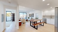 Located on Beverly Boulevard in Los Angeles, this modern 2-bedroom, 2-bathroom residence offers a thoughtfully designed open-concept layout finished with light hardwood flooring and crisp white walls throughout. The combined kitchen, dining, and living area forms the heart of the home, where a sleek kitchen features white shaker cabinetry, subway tile backsplash, and stainless steel appliances including a side-by-side refrigerator and electric range. A live-edge wood dining table anchors the dining area, which flows seamlessly into the bright living room. Large sliding glass doors flood the living space with natural light and open onto a spacious private balcony with sweeping views of the Los Angeles skyline and surrounding hills — ideal for outdoor lounging. The primary bedroom includes a ceiling fan, a sliding glass door to an outdoor area, and generous proportions, while both bedrooms are finished with the same warm hardwood flooring. The bathrooms are cleanly appointed with white vanities, large mirrors, and a tub-shower combination with sliding glass doors. A dedicated closet adds practical storage. Recessed lighting throughout enhances the bright, airy ambiance of this well-appointed Los Angeles residence.