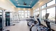 Located at 19770 Clay Rd in Katy, Texas, this well-appointed fitness center is a single-floor facility designed to support a comprehensive workout experience. The space is defined by its soaring ceilings with a distinctive teal-painted tray ceiling accent, recessed lighting, and a ceiling fan that keeps the environment comfortable during intense sessions. Large floor-to-ceiling windows line multiple walls, flooding the interior with natural light and offering open views of the surrounding community and outdoor amenities. The flooring throughout is a durable, cushioned exercise surface ideal for both cardio and strength training activities. The cardio zone is generously equipped with multiple treadmills, elliptical trainers, and a stationary bike, all positioned to take advantage of the bright, window-lined walls. The strength training area features a multi-functional cable machine, a squat and pull-up rack, adjustable benches, and a full rack of dumbbells ranging across various weights. Full-length mirrors are strategically placed to allow users to monitor form, and a large wall-mounted screen adds an entertainment element to the space. Decorative abstract wall art adds a modern aesthetic touch, contributing to an energizing yet polished atmosphere throughout the facility.
