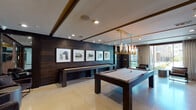 This stylish single-floor amenity space is designed for recreation and social engagement, blending contemporary aesthetics with premium entertainment features. The main game area is anchored by a dark-walnut pool table centered beneath a striking gold and glass cylindrical pendant chandelier, setting an upscale tone. Adjacent to it, a full-length shuffleboard table runs along a richly paneled dark wood accent wall, which also serves as a gallery backdrop for a curated series of four framed mixed-media artworks. Large-format light-toned tile flooring spans the entire space, offering a clean contrast to the warm, dark woodwork throughout. Recessed ceiling lighting and coffered ceiling details add architectural depth and dimension. A dedicated lounge zone features a wall-mounted flat-screen TV framed within a dark wood niche, complemented by upholstered bench seating with padded wall panels that lend a refined, comfortable feel. Floor-to-ceiling windows and a glass sliding door flood the space with natural light and provide views of an outdoor terrace. Entry is through a set of elegant dark-framed glass double doors, reinforcing the space's polished, modern character. The overall ambiance is sophisticated yet inviting, ideal for both casual gatherings and spirited game nights.