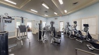 This single-story fitness center is designed to accommodate a wide range of exercise needs across several distinct zones, all unified by a cohesive aesthetic of soft blue walls, white decorative millwork, and coffered ceilings with recessed fluorescent lighting. Durable rubber flooring runs throughout, providing both safety and functionality across the facility.

The main strength training area is generously proportioned, housing a variety of Magnum-brand selectorized machines, cable systems, free weights, and a power rack with bench press. Full-length mirrors line the walls, enhancing the sense of space. An adjacent cardio zone features treadmills, ellipticals, and upright bikes positioned near windows that allow natural light to filter in.

A dedicated cycling studio accommodates a row of stationary spin bikes alongside wall-mounted mirrors and a mounted television. A separate multi-purpose exercise room provides open floor space with stored mats, medicine balls, and small fitness accessories, with interior windows overlooking an adjacent basketball court.

A charming children's play area features whimsical hand-painted murals, natural light from double windows, and age-appropriate toys and furnishings. Throughout the facility, white French-style doors connect the various zones, contributing to an open, airy, and welcoming atmosphere.