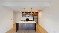This thoughtfully designed single-floor residence offers a clean, contemporary aesthetic with an open and functional layout. The kitchen serves as a central highlight, featuring rich dark-stained cabinetry, white quartz countertops, a full suite of stainless steel appliances including an electric range, built-in microwave, and refrigerator. A spacious center island with an integrated sink and pendant lighting above creates an inviting focal point that anchors the living area. Light hardwood-style flooring flows throughout the main living spaces, lending warmth and continuity to the interior. The living area leads to a private balcony through full-height sliding glass doors, which flood the space with natural light and offer open views of surrounding mature trees. The bedroom is generously proportioned, also benefiting from large sliding glass doors and plush carpeting underfoot. The bathroom is finished with light-toned tile flooring, a clean white vanity with an undermount sink, a large mirror, and a separate shower area. A dedicated utility closet accommodates a stacked in-unit washer and dryer, adding everyday convenience. Additional storage is available via a walk-in closet. Overall, the space presents a bright, well-appointed environment with modern fixtures and a practical, livable design.