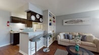 This single-floor residence offers a thoughtfully designed layout that maximizes both function and style. The open-concept living and kitchen area serves as the heart of the home, where rich hardwood flooring flows throughout. The kitchen is outfitted with dark espresso cabinetry, granite-style countertops, a double basin sink, a dishwasher, and a full-size refrigerator, complemented by a breakfast bar with modern bar stools — ideal for casual dining. A built-in white shelving unit elegantly divides the kitchen from the living room, which features a plush sofa, a glass-top coffee table, and large windows dressed with sheer curtains that invite natural light. The dedicated dining area accommodates a full dining table and chairs beneath a pendant light fixture. The bedroom is a serene retreat with carpeted flooring, a ceiling fan, natural light from a large window, and ample storage provided by a standout teal armoire. The full bathroom is well-appointed with dark cabinetry, granite-style countertops, a large vanity mirror, and hardwood-style flooring, completing this cohesive and comfortable living space.