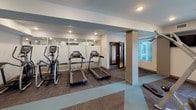 This fitness center is designed to support a comprehensive workout experience within a single-level layout. The space is divided into distinct functional zones, each dedicated to a different category of exercise. The main area features a free weights section with a full rack of dumbbells, adjustable weight benches, and a selection of medicine and stability balls stored on wall-mounted racks. Large mirrors line multiple walls, amplifying the sense of space and allowing for proper form checks during training.

The cardio zone is equipped with multiple treadmills and elliptical machines, positioned near floor-to-ceiling glass panels and windows that invite natural light and provide outdoor views. A dedicated cable and multi-station strength machine occupies an additional section, accompanied by a leg press bench for targeted resistance training.

An adjacent studio space is visible through interior glass walls, offering a separate area for cycling equipment and group or individual exercise. Recessed lighting throughout ensures consistent illumination across all zones. The flooring combines carpet tiles and rubberized stretching areas, providing both comfort and functionality. The overall design is clean, modern, and efficiently organized to accommodate a wide range of fitness activities.