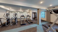 This fitness center is designed to support a comprehensive workout experience within a single-level layout. The space is divided into distinct functional zones, each dedicated to a different category of exercise. The main area features a free weights section with a full rack of dumbbells, adjustable weight benches, and a selection of medicine and stability balls stored on wall-mounted racks. Large mirrors line multiple walls, amplifying the sense of space and allowing for proper form checks during training.

The cardio zone is equipped with multiple treadmills and elliptical machines, positioned near floor-to-ceiling glass panels and windows that invite natural light and provide outdoor views. A dedicated cable and multi-station strength machine occupies an additional section, accompanied by a leg press bench for targeted resistance training.

An adjacent studio space is visible through interior glass walls, offering a separate area for cycling equipment and group or individual exercise. Recessed lighting throughout ensures consistent illumination across all zones. The flooring combines carpet tiles and rubberized stretching areas, providing both comfort and functionality. The overall design is clean, modern, and efficiently organized to accommodate a wide range of fitness activities.