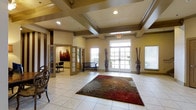 This single-floor space makes a strong first impression with its thoughtfully designed lobby and reception area. The entrance foyer features distinctive coffered ceilings with recessed lighting, creating a sense of architectural depth and grandeur. Light marble tile flooring extends throughout the main area, complemented by a bold, decorative area rug that anchors the welcoming space. Large glass-paned entry doors allow an abundance of natural light to flood the interior, enhancing the warm, neutral tones of the walls. Tall decorative vases flank the entrance, adding an elegant finishing touch. Adjacent to the main lobby, a secondary room is visible through open doorways, offering additional seating and wall art that contributes to the refined atmosphere.

The exterior covered porch is a standout feature, framed by substantial round columns set on brick bases and a herringbone-patterned brick floor. This outdoor space opens to sweeping views of a tranquil pond surrounded by manicured landscaping, mature trees, and neighboring buildings. Benches placed throughout the grounds invite residents or visitors to enjoy the serene outdoor setting. Together, the interior and exterior spaces convey a professional, polished environment that balances comfort with sophistication.
