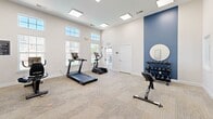 Located at 3737 Cusseta Rd in Columbus, Georgia, this fitness center offers a clean, modern environment designed to support a well-rounded workout routine. The space is characterized by tall ceilings and an abundance of natural light streaming through multiple large windows positioned along the walls, creating an open and energizing atmosphere. Recessed skylight-style ceiling panels provide additional overhead illumination, keeping the room bright and inviting throughout the day.

The interior features a neutral color palette of crisp whites accented by a bold navy blue feature wall, adding a contemporary and dynamic visual element. Durable, textured carpet tile flooring covers the workout area, offering both comfort and practicality underfoot.

The room is thoughtfully equipped with a range of cardio machines, including a treadmill, elliptical trainer, stationary bike, and recumbent bike. A tiered dumbbell rack with a full set of free weights and an adjustable workout bench provide strength training options. A wall-mounted smart mirror fitness display adds a modern, tech-forward touch for guided workouts.

Decorative artwork adorns the walls, contributing to an uplifting and polished aesthetic. Two access doors provide convenient entry and exit. Overall, this fitness center combines functionality with a welcoming design to support an active lifestyle.