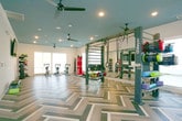 Located at 800 Crawford Street in Portsmouth, Virginia, this single-floor fitness center is a thoughtfully designed space that combines style with comprehensive workout functionality. The facility is immediately distinguished by its bold, multi-toned herringbone hardwood flooring, which runs throughout the entire space and creates a visually striking aesthetic. Tray ceilings painted in a deep teal-blue contrast elegantly with the crisp white walls, while recessed lighting and modern ceiling fans provide both illumination and ventilation throughout.

The gym is divided into distinct workout zones. A strength training area houses a full array of cable machines, weight benches, a squat rack, and free weights. Adjacent to this is a functional training zone equipped with power racks, gymnastic rings, and a heavy punching bag, ideal for dynamic workouts. A dedicated cardio section features treadmills, elliptical machines, and cycling bikes. A dumbbell rack and accessory equipment round out the offerings. Full-length mirrors are strategically placed to support form monitoring during training.

Large windows fitted with sheer roller shades allow abundant natural light to fill the space while offering views of the surrounding Portsmouth neighborhood. A water fountain station and wall-mounted television add convenience and comfort. Glass doors provide access to an outdoor area, enhancing the open, airy atmosphere of this well-appointed fitness facility.