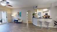 This single-story residence is designed with an open, flowing layout that connects the main living areas seamlessly. Wood-look flooring in warm gray tones runs throughout, providing a cohesive and modern aesthetic across every room.

The main living area is spacious and airy, featuring a ceiling fan and a sliding glass door that invites natural light while offering access to an outdoor patio. A distinctive arched doorway serves as an architectural accent, connecting the living space to the rest of the unit and adding a classic design touch.

The kitchen is well-appointed with crisp white cabinetry, dark stone-look countertops, and stainless steel appliances including a range, microwave, and refrigerator. A breakfast bar with modern bar seating provides a casual dining option and opens directly to the adjacent dining area, which is illuminated by a pendant light overhead. A louvered closet door in the dining area offers convenient storage.

Additional rooms include a generously sized bedroom with its own ceiling fan, a window allowing natural light, and direct access back to the main living area through an arched passage. The bathroom features a full tub and shower combination. Throughout, neutral wall tones and clean finishes create a bright, welcoming ambiance.