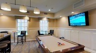This single-floor amenity space is thoughtfully designed as a social and recreational retreat. At its center sits a full-sized pool table with a cream felt surface, positioned beneath stylish drum pendant lights that provide focused, warm illumination. The room's design strikes a balance between comfort and sophistication, featuring wainscoting along the lower walls and warm neutral tones throughout that lend a refined, welcoming atmosphere.

A wall-mounted flat-screen television is positioned prominently, making the space equally suited for watching live sports or entertainment. Natural light fills the room through multiple large double-hung windows dressed with decorative valances, offering views of the brick exterior surroundings. Recessed ceiling lights complement the pendant fixtures, ensuring the room is well-lit throughout.

Seating is provided by sleek black leather bar-height chairs arranged near a tall bistro table beneath the windows, creating an ideal spot for casual conversation. The patterned carpet underfoot adds texture and visual interest, tying the room's classic design elements together. Overall, this space offers a versatile and inviting environment well-suited for recreational activities and social interaction.