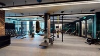 This fitness center is designed with a thoughtful industrial-modern aesthetic, combining exposed brick accent walls, black steel structural elements, and deep teal wall finishes to create a stylish yet functional environment. Linear LED lighting runs along the ceiling throughout, providing consistent, even illumination across the workout floor.

The main exercise area is generously proportioned, accommodating a wide range of equipment including treadmills, rowing machines, stair climbers, stationary bikes, and a hanging heavy bag. A multi-functional cable and suspension training rig anchors the central zone, while a dedicated free weights station lines the brick wall, complete with a full dumbbell rack and adjustable bench. Floor-to-ceiling windows along one side of the main floor flood the space with natural light and offer views of the surrounding exterior. Durable rubber flooring covers the entire workout area.

A separate, more intimate studio room features warm hardwood floors, full-wall mirrors with a ballet barre, a wall-mounted television, and an array of accessories including stability balls and kettlebells—well-suited for yoga, barre, or functional training sessions. Adjacent to the main gym, a glass-enclosed retail area provides convenient access to beverages and snacks.