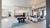 This contemporary single-floor clubhouse is designed to impress, offering a wide range of amenity spaces wrapped in a cohesive modern aesthetic. The main lounge area serves as the heart of the space, featuring soaring ceilings, large floor-to-ceiling sliding glass doors that invite abundant natural light, and an eye-catching sputnik chandelier overhead. A full-size billiards table anchors the room, complemented by plush seating arrangements on patterned area rugs.

The fully equipped kitchen is a standout feature, showcasing a large island with a stainless steel countertop and sleek black barstools, warm walnut cabinetry, a statement mosaic tile backsplash, and striking geometric diamond pendant lights. A full-size stainless refrigerator and open display shelving complete the space.

The lounge seating area offers a more intimate setting with textured accent walls, a wall-mounted flat-screen TV, and modern armchairs arranged around a glass coffee table, with direct views to an outdoor patio area.

A spacious waiting or lobby area features dramatic double-height windows, a bold navy accent wall, and a chevron art installation, creating a striking first impression. The business center includes workstations and computer terminals, while a dedicated conference room provides a professional setting with a large table, ergonomic seating, and stylish pendant lighting. The entry foyer features rich textured wall panels and elegant wood cabinetry.