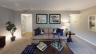 This thoughtfully designed single-floor residence offers a cohesive blend of comfort and functionality across its well-proportioned rooms. The living room serves as the heart of the home, featuring plush carpeting, a sliding glass door that opens to a private balcony with lush outdoor views, and generous space for both relaxing and entertaining. Adjacent to the living area, the dining room benefits from a large window that fills the space with natural light, complemented by a pendant light fixture overhead and hardwood-style flooring. The kitchen is equipped with white shaker cabinetry, granite countertops, a classic subway tile backsplash, and a full suite of stainless steel appliances including a gas range, dishwasher, and built-in microwave. Two well-sized bedrooms feature soft carpeting, ample natural light from windows with decorative curtains, and a calm, neutral color palette. A versatile bonus room functions as a dedicated home office, complete with built-in shelving and a comfortable seating area. Two full bathrooms each include a tub-shower combination with tile flooring and clean white vanities. Throughout the residence, a consistent neutral gray tone and coordinated blue accents create a refined and welcoming atmosphere.