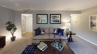 This thoughtfully designed single-floor residence offers a cohesive blend of comfort and functionality across its well-proportioned rooms. The living room serves as the heart of the home, featuring plush carpeting, a sliding glass door that opens to a private balcony with lush outdoor views, and generous space for both relaxing and entertaining. Adjacent to the living area, the dining room benefits from a large window that fills the space with natural light, complemented by a pendant light fixture overhead and hardwood-style flooring. The kitchen is equipped with white shaker cabinetry, granite countertops, a classic subway tile backsplash, and a full suite of stainless steel appliances including a gas range, dishwasher, and built-in microwave. Two well-sized bedrooms feature soft carpeting, ample natural light from windows with decorative curtains, and a calm, neutral color palette. A versatile bonus room functions as a dedicated home office, complete with built-in shelving and a comfortable seating area. Two full bathrooms each include a tub-shower combination with tile flooring and clean white vanities. Throughout the residence, a consistent neutral gray tone and coordinated blue accents create a refined and welcoming atmosphere.