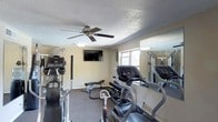 This fitness room is a dedicated exercise space designed to support a well-rounded workout routine. The room is outfitted with a selection of cardio equipment, including treadmills and an elliptical machine, positioned to make efficient use of the available floor space. A set of free weights and a adjustable workout bench provide options for strength training, making the room suitable for both cardio and resistance-based exercise.

A wall-mounted flat-screen television is positioned for easy viewing during workouts, adding an entertainment element to the space. The ceiling features a textured finish and is equipped with a ceiling fan, which aids in air circulation during exercise sessions. Flush-mounted light fixtures supplement the natural light that enters through a window overlooking the exterior of the building.

Full-length mirrors are installed along one wall, serving both a functional and spatial purpose by reflecting light and giving the room a more open feel. The flooring is covered in dark, durable carpeting suited for gym use. The overall layout is practical and functional, with equipment arranged to allow clear movement throughout the space.
