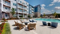Located at 4129 Falcon Pkwy in Oakwood, Georgia, this outdoor amenity space is the centerpiece of a modern multi-story residential community. The architecture blends blue-paneled siding, classic brick masonry, and white trim across multiple building wings that frame the courtyard, creating an enclosed and cohesive environment. Private balconies extend from upper floors, offering elevated views of the pool area below.

The expansive resort-style swimming pool is the focal point of the outdoor space, featuring clear blue water, built-in entry steps, and a shallow wading ledge along one edge. Surrounding the pool is a generously sized concrete sundeck furnished with rows of sleek charcoal lounge chairs and side tables, ideal for relaxation. Umbrella-shaded seating clusters and woven lounge chairs with cushions provide additional gathering areas for social interaction.

Thoughtful landscaping softens the hardscape with neatly trimmed shrubs, young trees, and planted garden beds along the building perimeter, adding greenery and visual texture. Fire bowl accents are positioned throughout the deck, suggesting year-round usability of the outdoor space. The courtyard benefits from abundant natural light and open sky views, enhancing the airy, resort-like atmosphere of this well-appointed community amenity in Oakwood, GA.