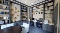 This thoughtfully designed office space combines functionality with refined aesthetics. The room is anchored by floor-to-ceiling built-in shelving units finished in a sophisticated charcoal gray, providing ample display and storage space. The shelves are tastefully styled with decorative wooden bowls, ceramic vases, woven baskets, and lush green plants, adding warmth and character to the space.

A built-in workstation runs along the back wall, comfortably accommodating two computer workstations, each equipped with monitors and ergonomic task chairs. The countertop surface is clean and functional, making it ideal for focused work. Cabinetry beneath the counters maintains the cohesive charcoal gray palette, offering additional storage.

The walls are finished in a soft, neutral cream tone that contrasts elegantly with the dark millwork and trim. A row of decorative wooden slice wall art adorns one side wall, contributing an organic, artistic touch. The patterned blue-gray carpeting adds texture underfoot while complementing the overall color scheme.

A small seating area near the front includes light gray upholstered chairs and a dark round table, offering a casual space for meetings or informal conversation. Recessed ceiling lighting ensures the room is well-lit throughout, creating a bright, welcoming, and professional environment.
