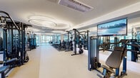 Located at 377 Crane St in Orange, NJ, this single-floor fitness center offers a spacious, thoughtfully designed workout facility with a sleek, modern aesthetic. The open layout is anchored by a striking ceiling treatment of large, overlapping circular LED light fixtures that cast a bright, even glow throughout the space, complementing the light-toned flooring and white walls to create an energetic yet clean atmosphere.

The main workout floor is comprehensively equipped with a wide array of strength training machines, including cable systems and multi-function units, alongside a well-stocked free weights section featuring a full dumbbell rack and adjustable benches. Floor-to-ceiling mirrors line the walls, amplifying the sense of space and allowing for proper form checks during exercise. Large windows along the perimeter invite abundant natural light and outdoor views, further enhancing the open feel of the facility.

A dedicated cardio zone houses a row of treadmills and stair climbers, positioned near the windows to take advantage of natural light. A polished cylindrical mirror column serves as a distinctive architectural accent in this area. A separate spin room, enclosed by sleek full-height glass partitions with frosted detailing, provides a private space for cycling workouts and is equipped with stationary bikes and a wall-mounted television. The facility's design balances functionality with a premium, upscale ambiance throughout.
